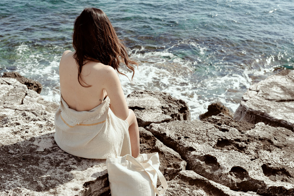 Roberta seduta sulla scogliera fronte mare con addosso il telo mare in lino e canapa